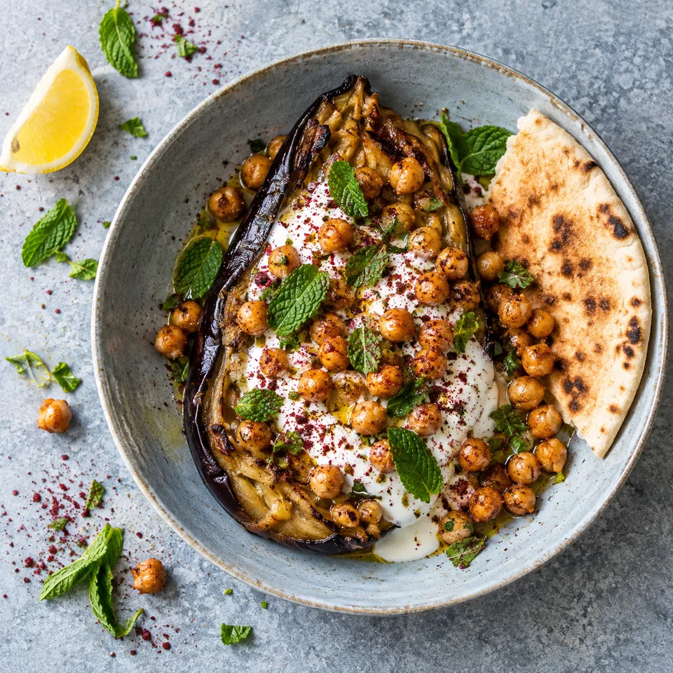 Aubergines grillées, yaourt à l’ail et pois chiches : aubergine fondante nappée de yaourt blanc, pois chiches dorés, sumac rouge et menthe verte sur assiette bleutée.