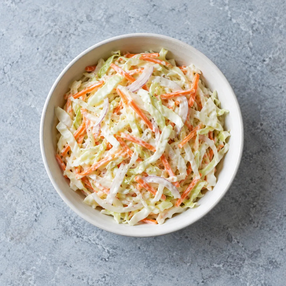 Portion de coleslaw croquant dans un bol blanc : chou blanc râpé, carotte orange et oignon enrobés d'une sauce blanche crémeuse.