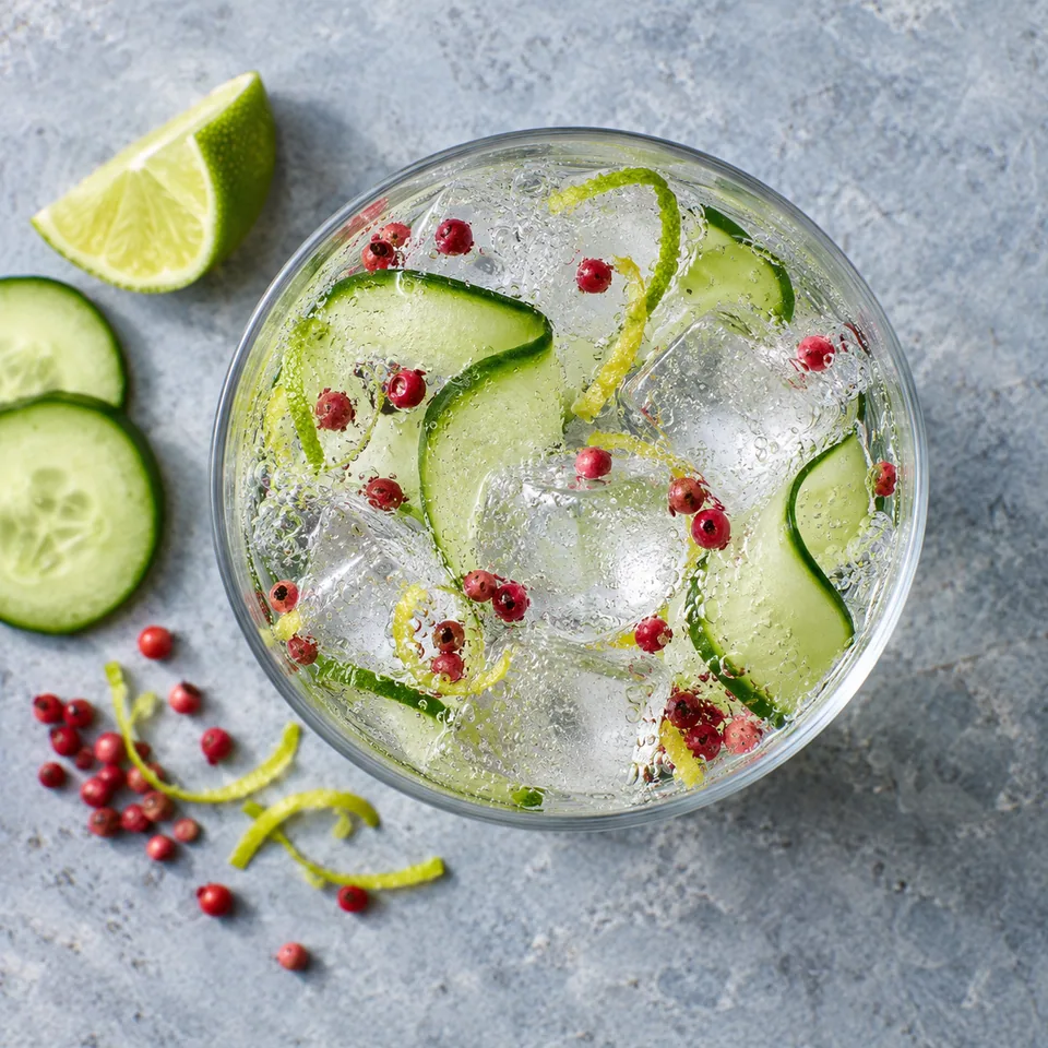 Gin tonic au concombre et au poivre rose dans un grand verre transparent avec glaçons, rubans de concombre et zestes.