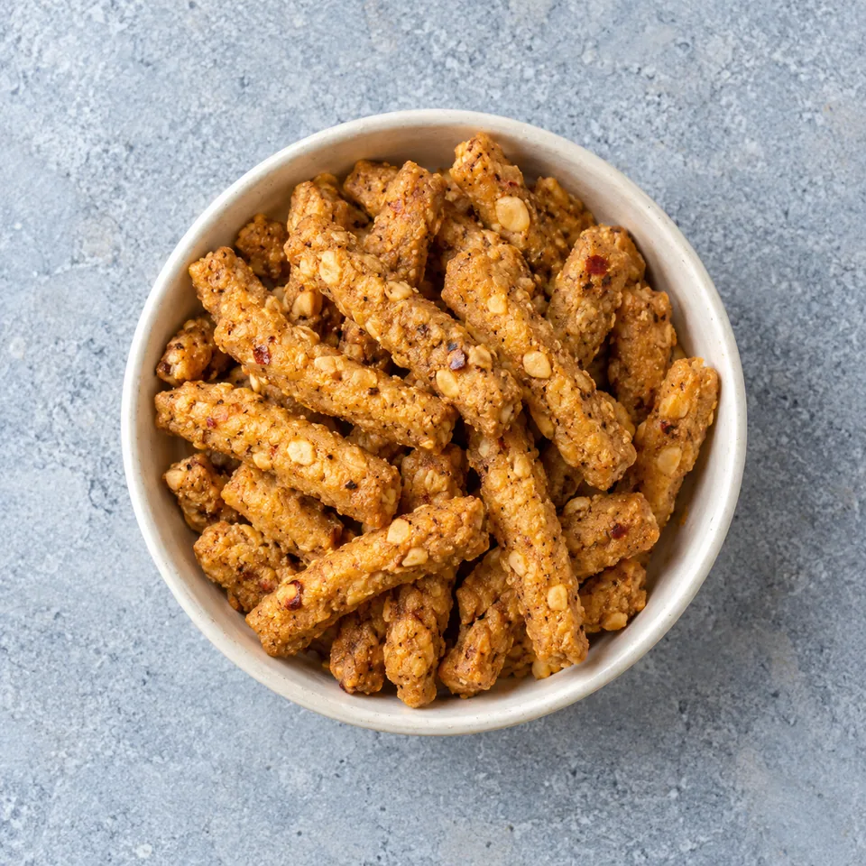 Kuli kuli au four : bâtonnets de cacahuètes épicés dorés et croustillants dans un petit bol blanc cassé.
