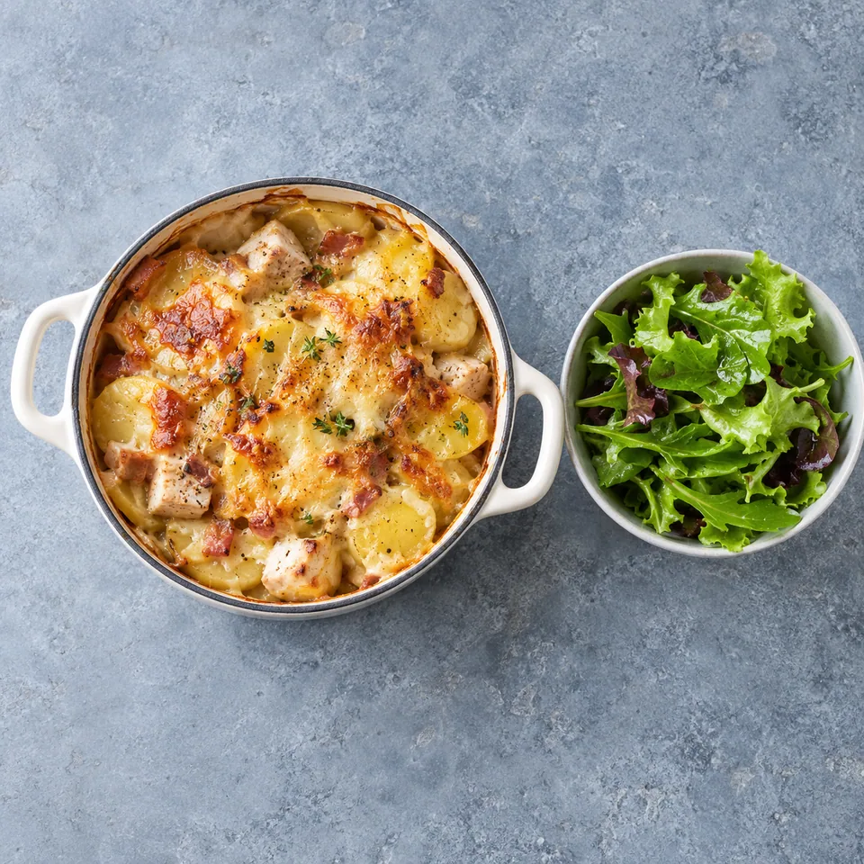 Portion de tartiflette au comté et à la dinde dans une cocotte blanche, gratinée dorée, accompagnée de salade verte.